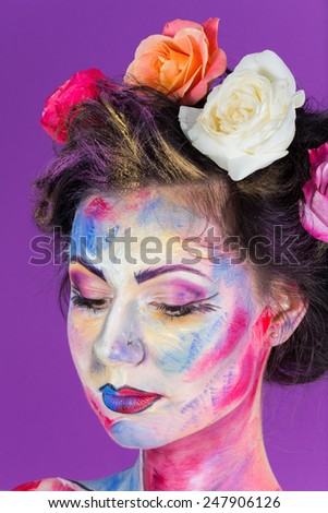 Beauty model on a purple background. Creative floral make-up on the face of the model. Multi-colored roses in her hair. Colorful makeup. Art make-up face, lips, eyes and hair.