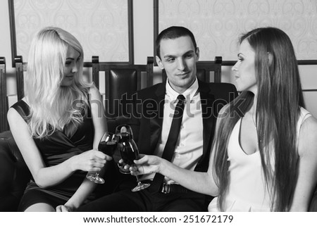 A man holding a glass of wine. Two girls standing in the background and drink wine. A man in a business suit and two pretty girls in the room. The concept of relationships between people.