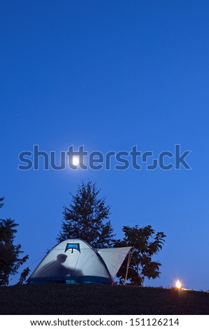 Camping in tent with human shadow and campfire, moon and stars on the clear blue sky