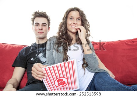 Close up portrait of young couple sitting together on a sofa at home watching television, joyfully smiling eating pop corn enjoying a night in together. Home lifestyle and entertainment technology.