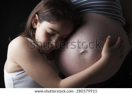 Beautiful little woman girl hugging pregnant mother\'s belly against black background.