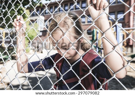 A sad little girl at school playground with fence on front of is fence.