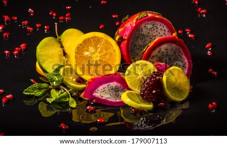 Colorful juicy assortment of sliced tropical fruits (purple dragon fruit, orange, lemon, pomegranate seeds, green apple and mint), on a wet black table with splashes of water