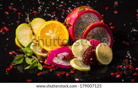 Colorful juicy assortment of sliced tropical fruits (purple dragon fruit, orange, lemon, pomegranate seeds, green apple and mint), on a wet black table with splashes of water