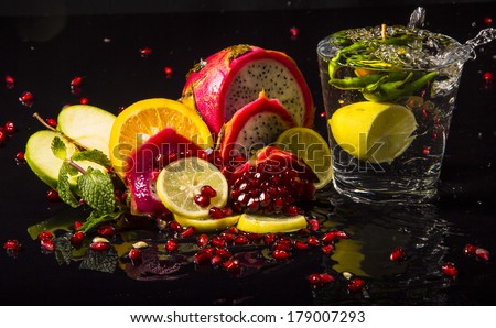 Colorful juicy assortment of sliced tropical fruits (purple dragon fruit, orange, lemon, pomegranate seeds, green apple and mint), on a wet black table, with a glass of transparent water
