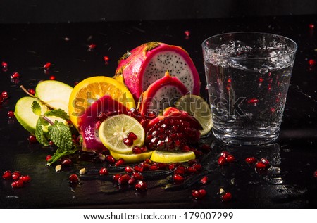 Colorful juicy assortment of sliced tropical fruits (purple dragon fruit, orange, lemon, pomegranate seeds, green apple and mint), on a wet black table with a glass of transparent water