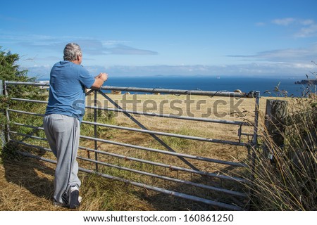 A man resting on a farm gate gazes out over welsh coastal field with the sea in the distance