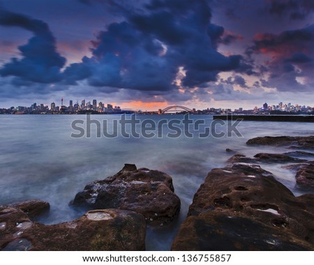 Sydney city CBD and harbour bridge at sunset windy cloudy weather blurred clouds dramatic sun light sandstone rocks on foreground with cityline and harbour water