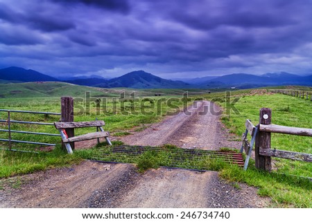 Rural agricultural property dividing fence, grid and gate on unsealed road through property in stormy valley with green grazing grass surrounded by hilly mountains