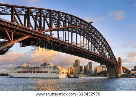 Oversized ocean cruise liner passing by under the Sydney Harbour Bridge at sunset backgrounded by city skyscrapers