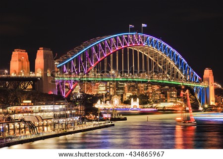 Brightly illuminated arch side of landmark Sydney Harbour Bridge after sunset reflecting lights in blurred still waters of bay.