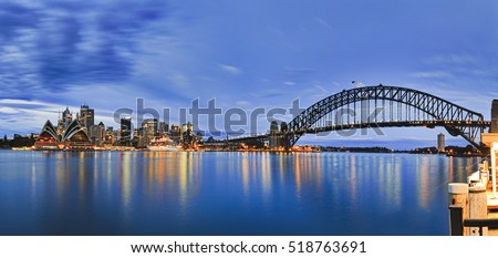 Landmarks of Sydney city CBD across Harbour with bridge, circular quay, passenger terminal and downtown skyscrapers to Kirribilli pier at blue cloudy sunrise.