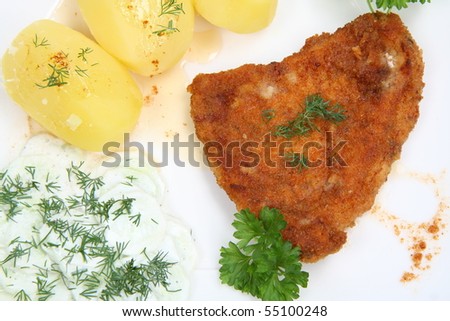 Pork chop (covered in batter and breadcrumbs), potatoes and cucumber salad decorated with dill and parsley on a plate on white background