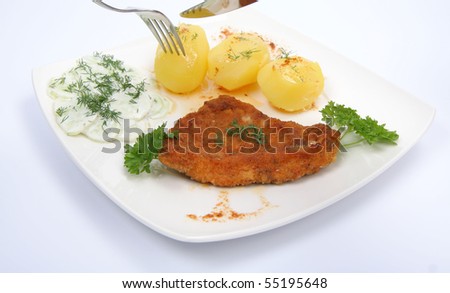 Pork chop (covered in batter and breadcrumbs), potatoes - being cut -and cucumber salad decorated with dill and parsley on a plate on white background