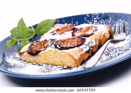 Piece of Plum Pie covered with powder sugar on a blue plate decorated with a mint twig and  a fork