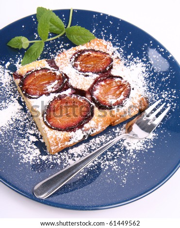 Piece of Plum Pie covered with powder sugar on a blue plate decorated with a mint twig