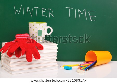 Books , colored pencil in yellow glass and winter gloves on green chalkboard. / End of school. Winter break