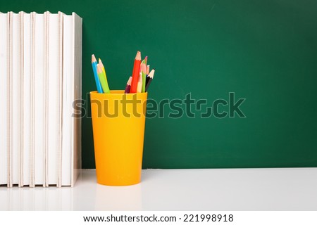 White books, colorful used pens in yellow glass on white desk against green chalkboard with copy-space
