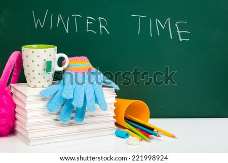 Books, winter gloves, cup of tea and used pens on white desk against green chalkboard. Winter time. Winter break