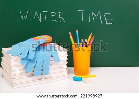 Books, winter gloves,  used pens in yellow cup on white desk against green chalkboard. Winter time. Winter break