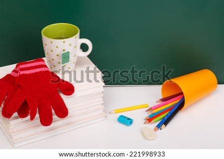 Books, winter gloves,  used pens in yellow cup on white desk against green chalkboard. Winter time. Winter break