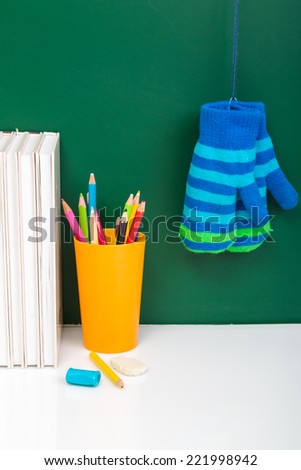 Books, winter gloves, used pens in yellow cup on white desk against green chalkboard. Winter time. Winter break Front view.