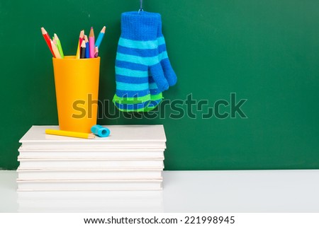 Books, winter gloves, used pens in yellow cup on white desk against green chalkboard with copy space Winter time. Winter break Front view.