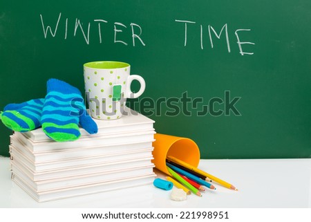 Books, winter gloves, cup of tea and used pens on white desk against green chalkboard. Winter time. Winter break