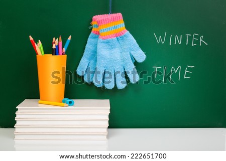White books, colorful used pens in yellow glass and winter gloves on white desk against green chalkboard with copy-space