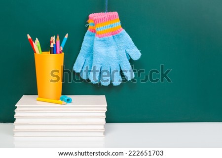White books, colorful used pens in yellow glass and winter gloves on white desk against green chalkboard with copy-space
