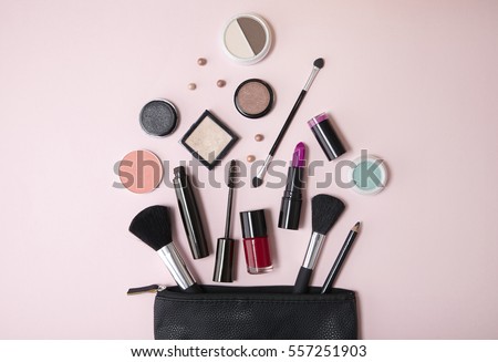 Overhead view of a black leather make up bag, with cosmetic beauty products spilling out on to a pastel pink background