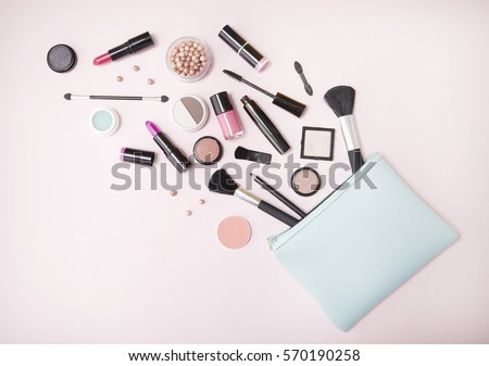 A blue makeup bag with cosmetic beauty products spilling out on to a pastel pink background