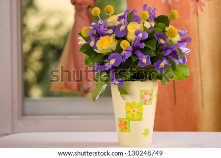 Yellow and lavender flowers in a vase next to a window with orange curtain