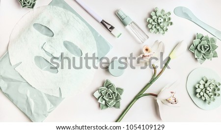 Skin care flat lay with facial sheet mask, mist spray bottle , succulents and orchid flowers on white desktop background, top view. Beauty spa and wellness concept