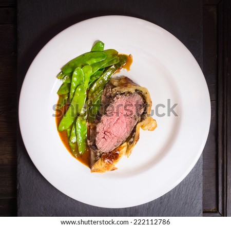 Beef Wellington with green peas and sauce  on dark wooden table, top view