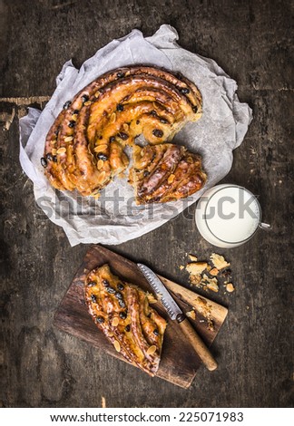 Sweet Braided cake with raisins and almonds cut to pieces  in white paper , cup of milk on dark wooden background