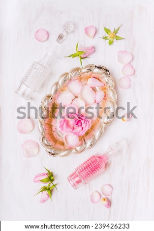 Silver bowl with roses in water,  bottle and buds and petals of roses on white wooden background, top view