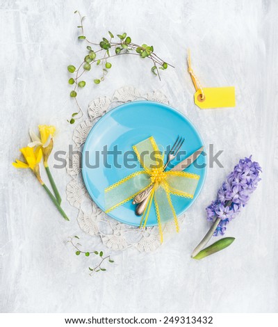 Blue plate with knife,fork, spring flowers and yellow ribbon decoration on gray wooden background, top view
