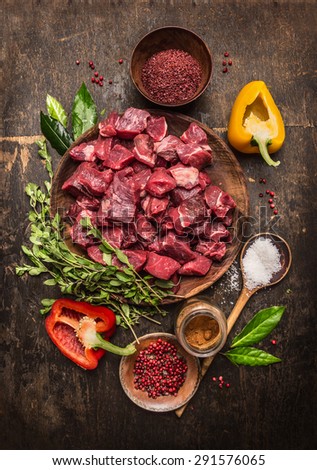 Raw  uncooked meat sliced in cubes with fresh herbs, vegetables and spices on rustic wooden background, ingredients for beef stew recipe, top view