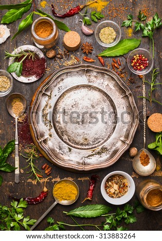 Colorful spices and herbs with spoon around empty vintage metal plate on rustic wooden background, top view