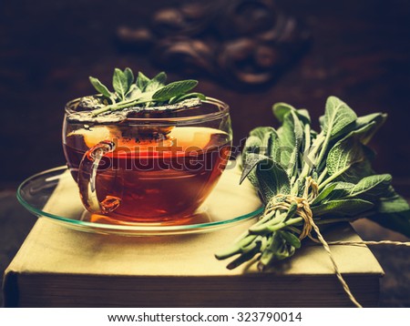 Herbal tea made from sage in glass cup standing on books, nearby lies a bundle of sage over dark wooden background. Retro toned.