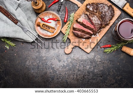 Roasted sliced grill steak on wooden cutting board with wine, seasoning and meat fork on dark vintage metal background, top view, border