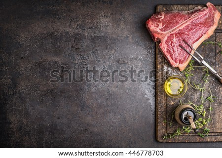 Raw T-bone Steak for grill or BBQ with meat fork and flavoring on aged cutting board and dark rustic metal background, top view, border