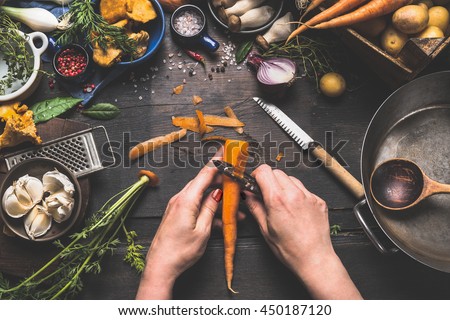 Female woman hands peeling carrots on dark wooden kitchen table with vegetables cooking ingredients, spoon and tools, top view
