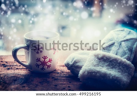 Snowflakes Mug with hot beverage and knitting mittens on frost window sill at winter snow nature background