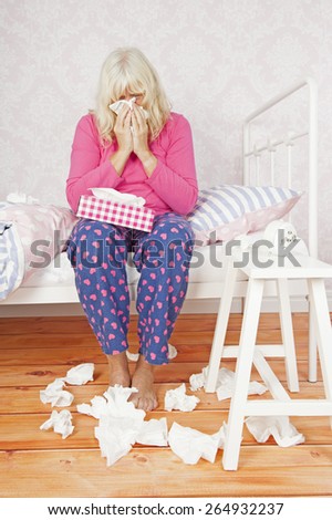 Sad female with pink pajama and tissues sitting on bed blowing her nose