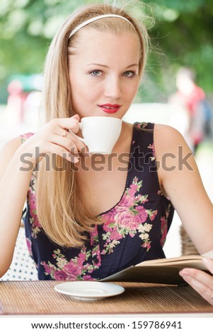beautiful girl drinks coffee and reads the book in cafe on the street in the summer