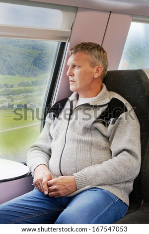 mature man in a gray cardigan sits in a car of a train and looks out of the window
