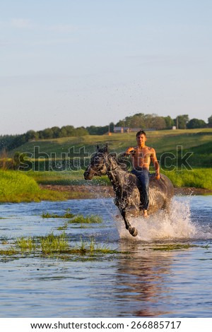 young man with a naked torso jumps astride a horse through the river