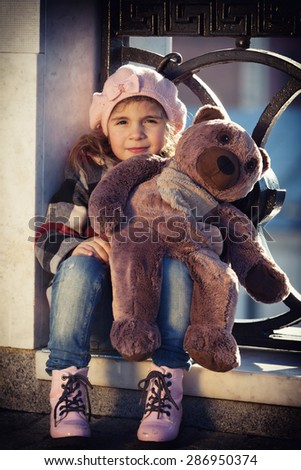 little girl in a pink beret sits at a fencing of the bridge and holds a toy bear in hand. Art processing: toning, vignetting